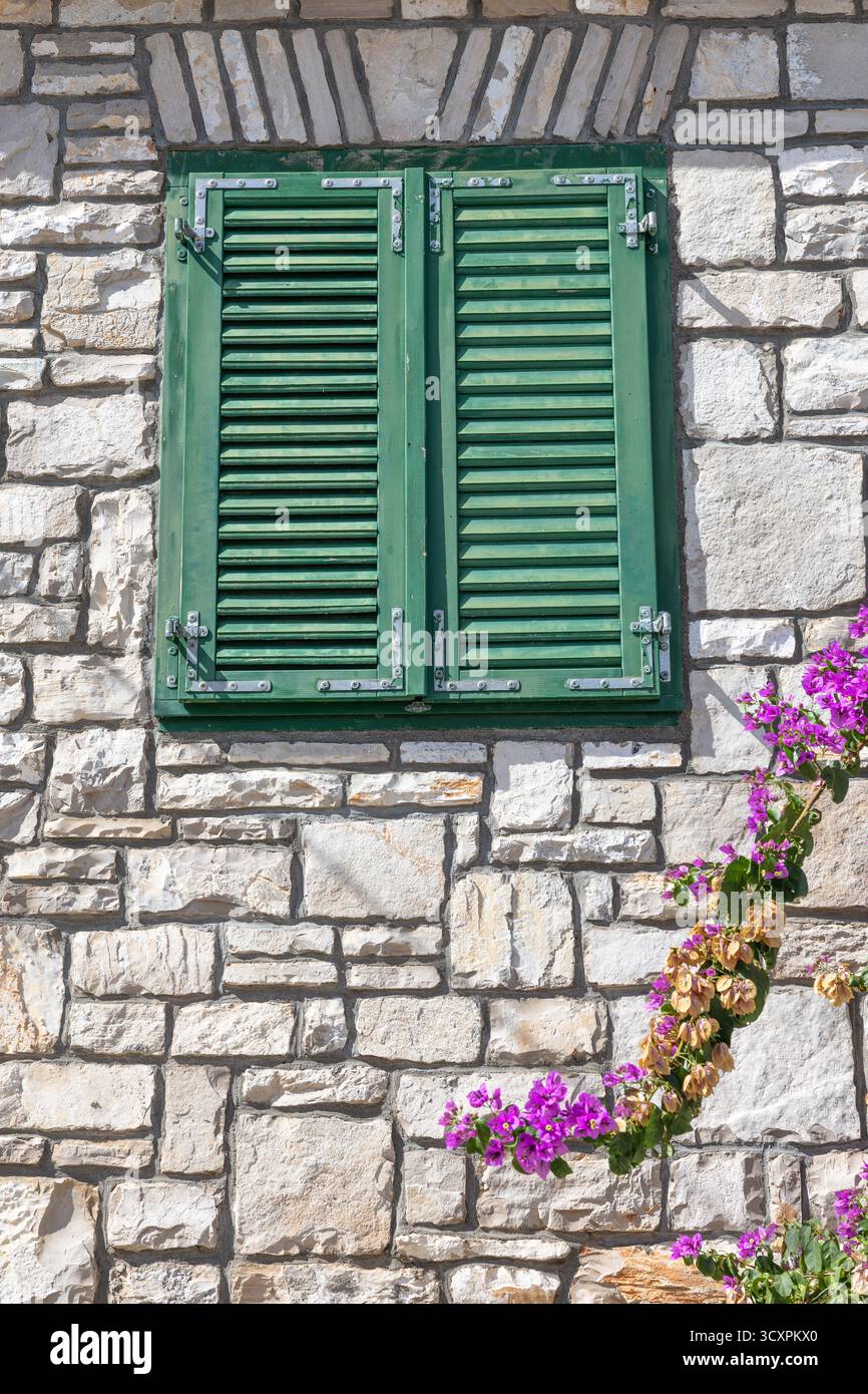 Una casa rustica in pietra con persiane verdi intemprate si trova accanto alle vivaci bouganville in piena fioritura sotto un cielo blu chiaro. Affascinante, caldo e immaginario Foto Stock
