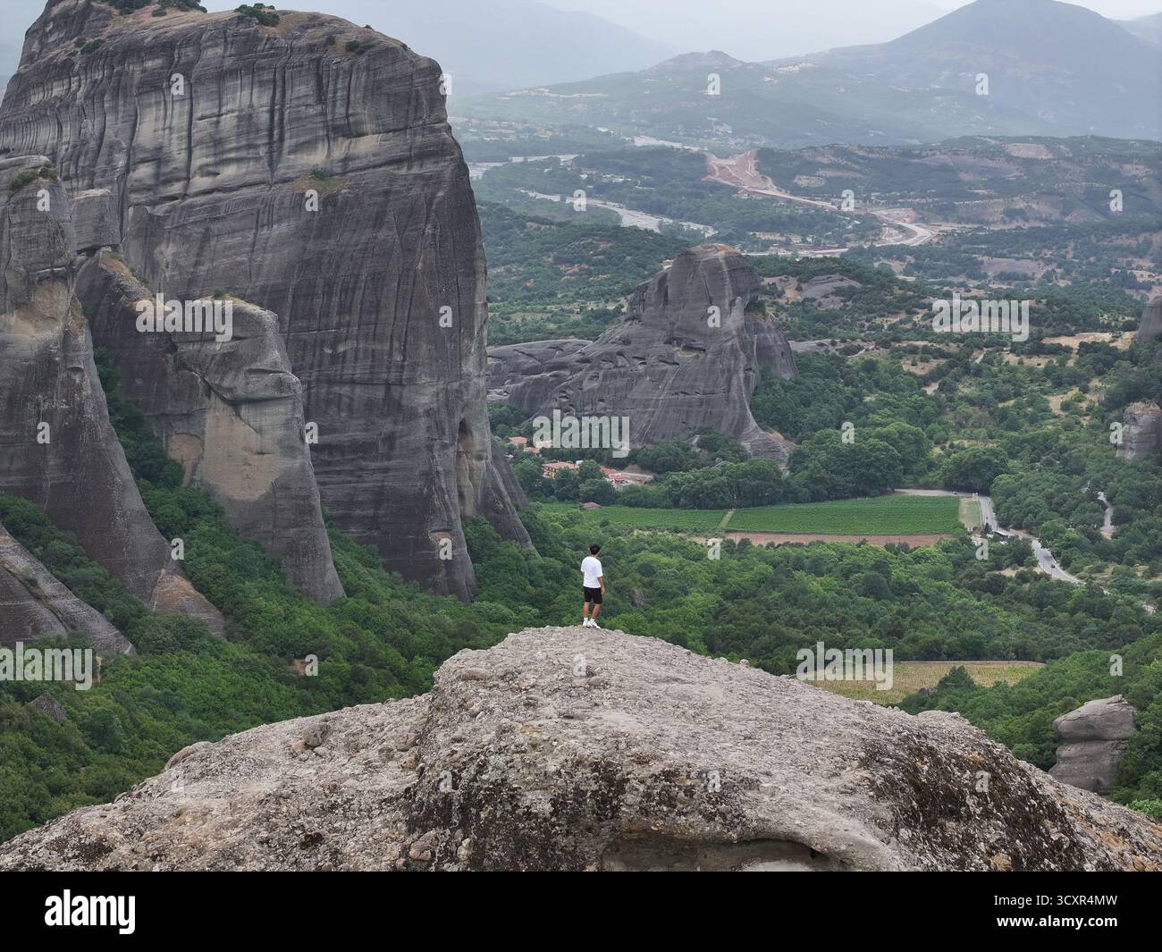 Uomo in piedi su una grande roccia, che si affaccia sul vasto e suggestivo paesaggio di meteora, grecia, con torreggianti formazioni rocciose, verdi vallate, Foto Stock