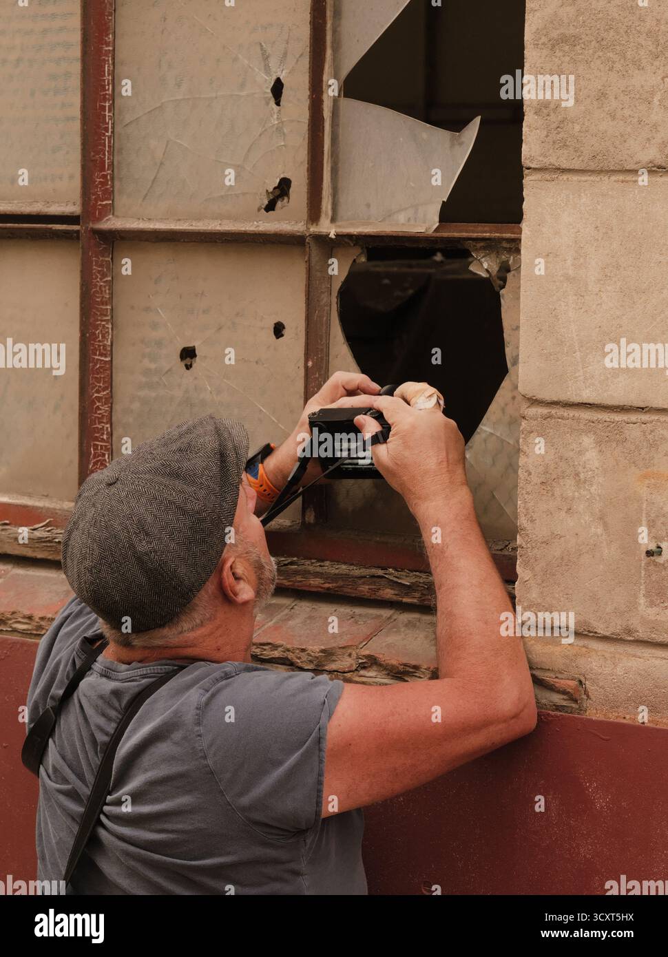 Fotografo con cappuccio piatto che cattura una scena di decadimento urbano attraverso una finestra rotta; grintoso, malinconico, riflettente. Foto Stock