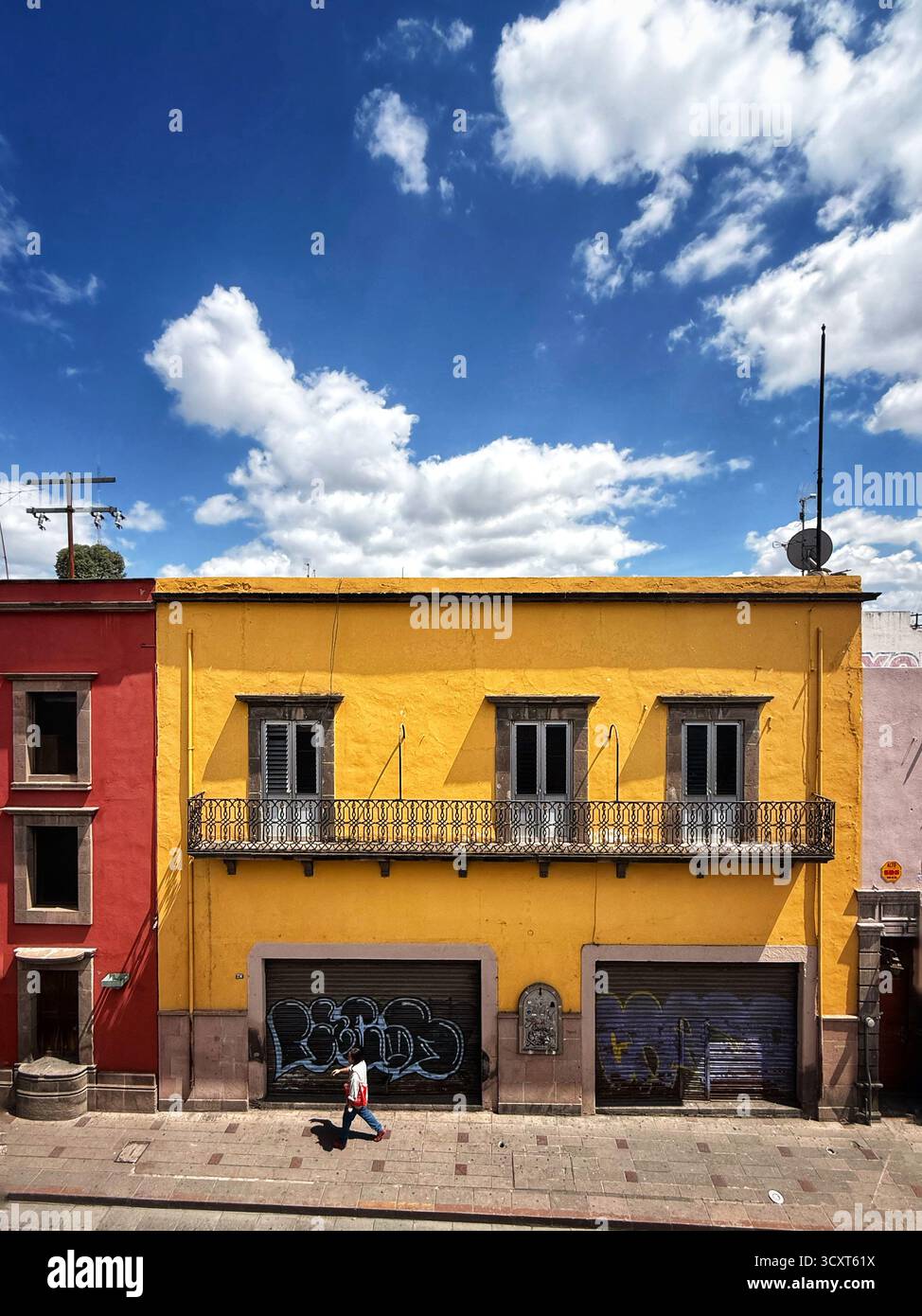 Vista colorata della strada con un edificio giallo vibrante e una persona che cammina sotto un cielo blu luminoso con nuvole sparse Foto Stock