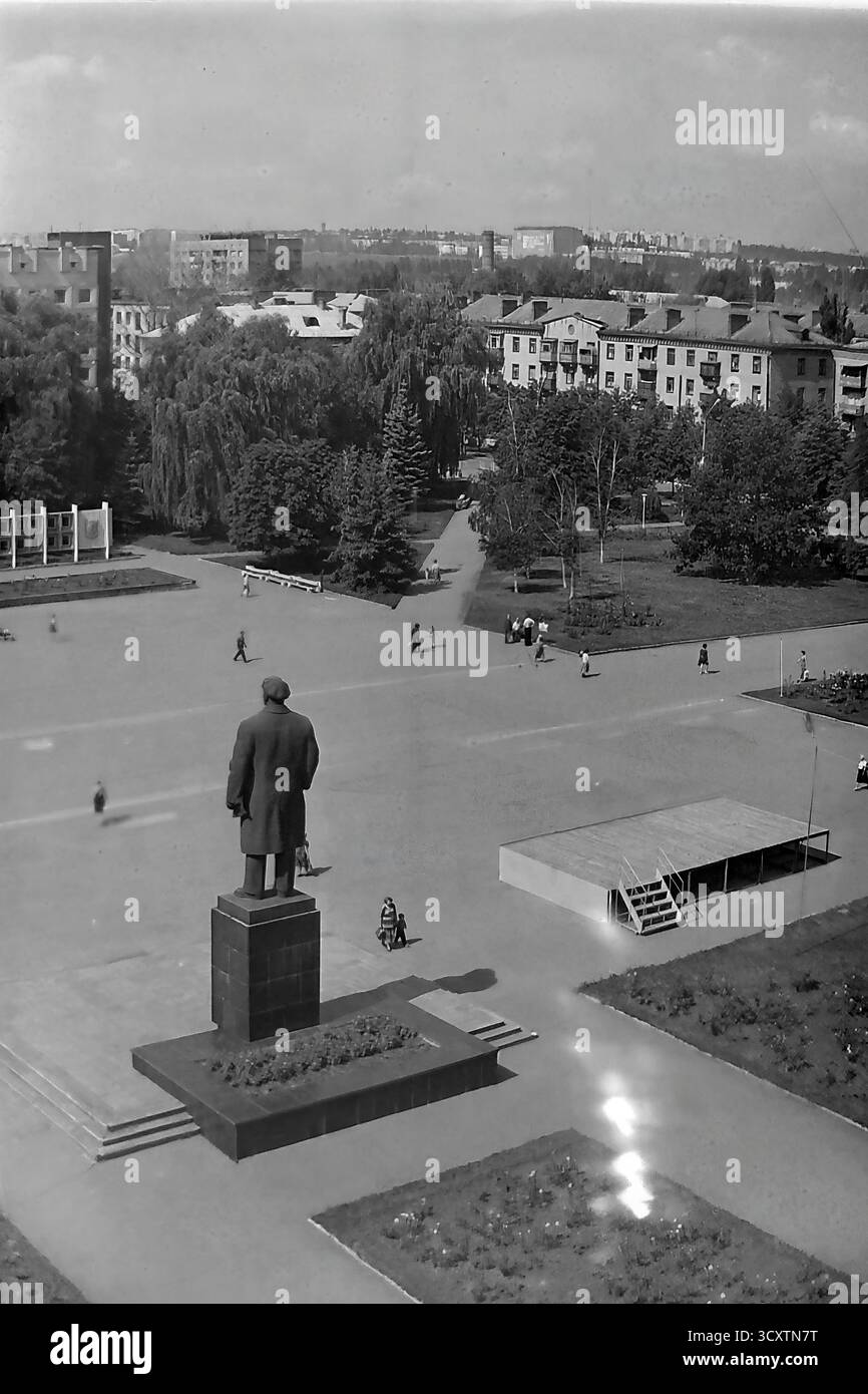Una foto d'archivio ad alto angolo degli anni '1980 cattura una vista di Piazza della Rivoluzione d'ottobre (ora Piazza Soborna) a Sloviansk. L'iconico monumento di Lenin è visibile da dietro, affacciato sul vasto spazio pubblico dove è stato eretto un palco temporaneo in legno per un evento. I pedoni, tra cui una donna con un passeggino, potranno godersi una giornata tranquilla nella piazza e nel parco adiacente. Questa scena è un'istantanea caratteristica della vita pubblica e degli eventi della comunità nell'era sovietica, una commovente testimonianza di un pacifico Donba prima della guerra Foto Stock