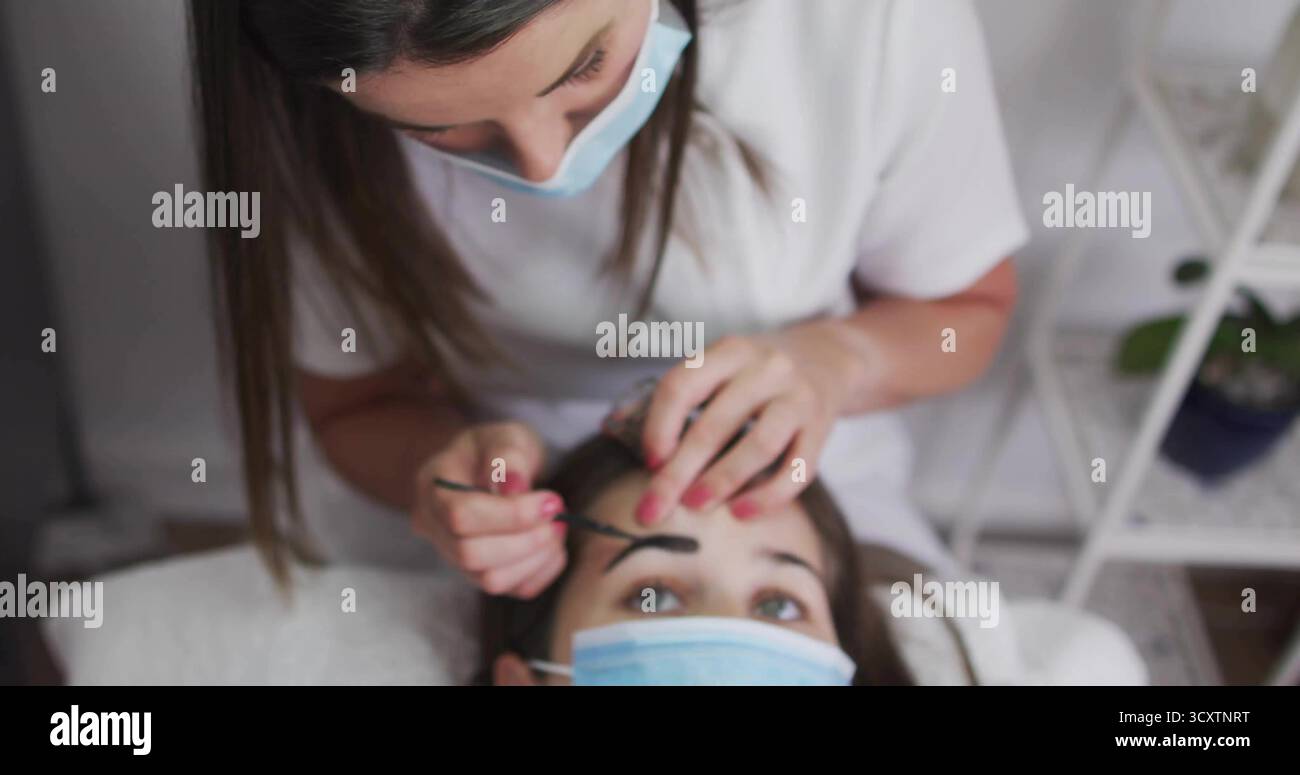 Estetista mascherato che applica siero sopracciglia al cliente su un letto bianco nella camera spa, con spazzola Foto Stock