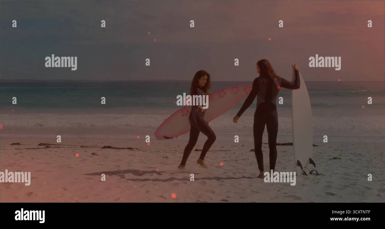 Tenendo in mano tavole da surf rosa e bianche, una coppia di fronte all'altra su una spiaggia sabbiosa, mute a tutta lunghezza Foto Stock