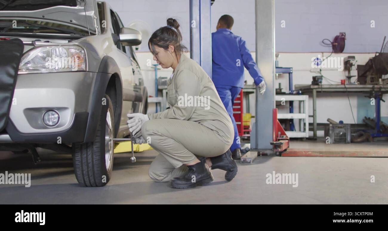 Accovacciato meccanico femmina che rimuove i dadi di fissaggio sul pavimento dell'officina sotto il sollevatore idraulico, con una chiave di manovra Foto Stock