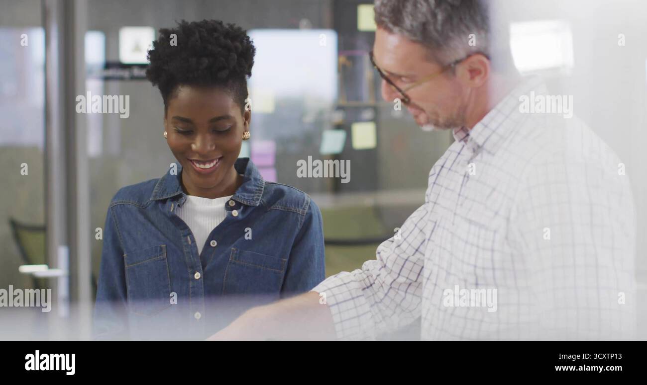 Uomo pendente che punta alla scrivania accanto al sorridente collaboratore in denim in ufficio, con appunti di separazione in vetro Foto Stock