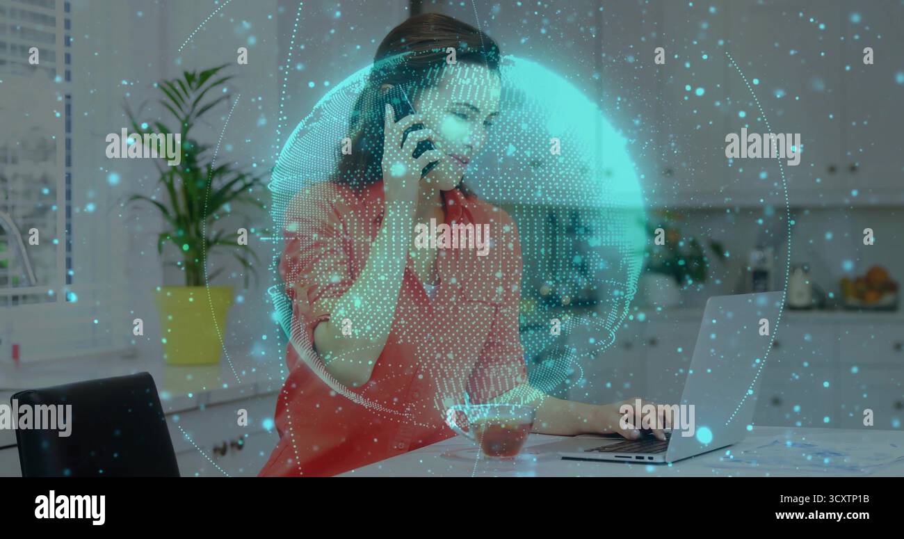 Donna parlante in camicetta rossa che utilizza lo smartphone mentre digita il laptop in cucina, con sovrapposizione sfera dati Foto Stock
