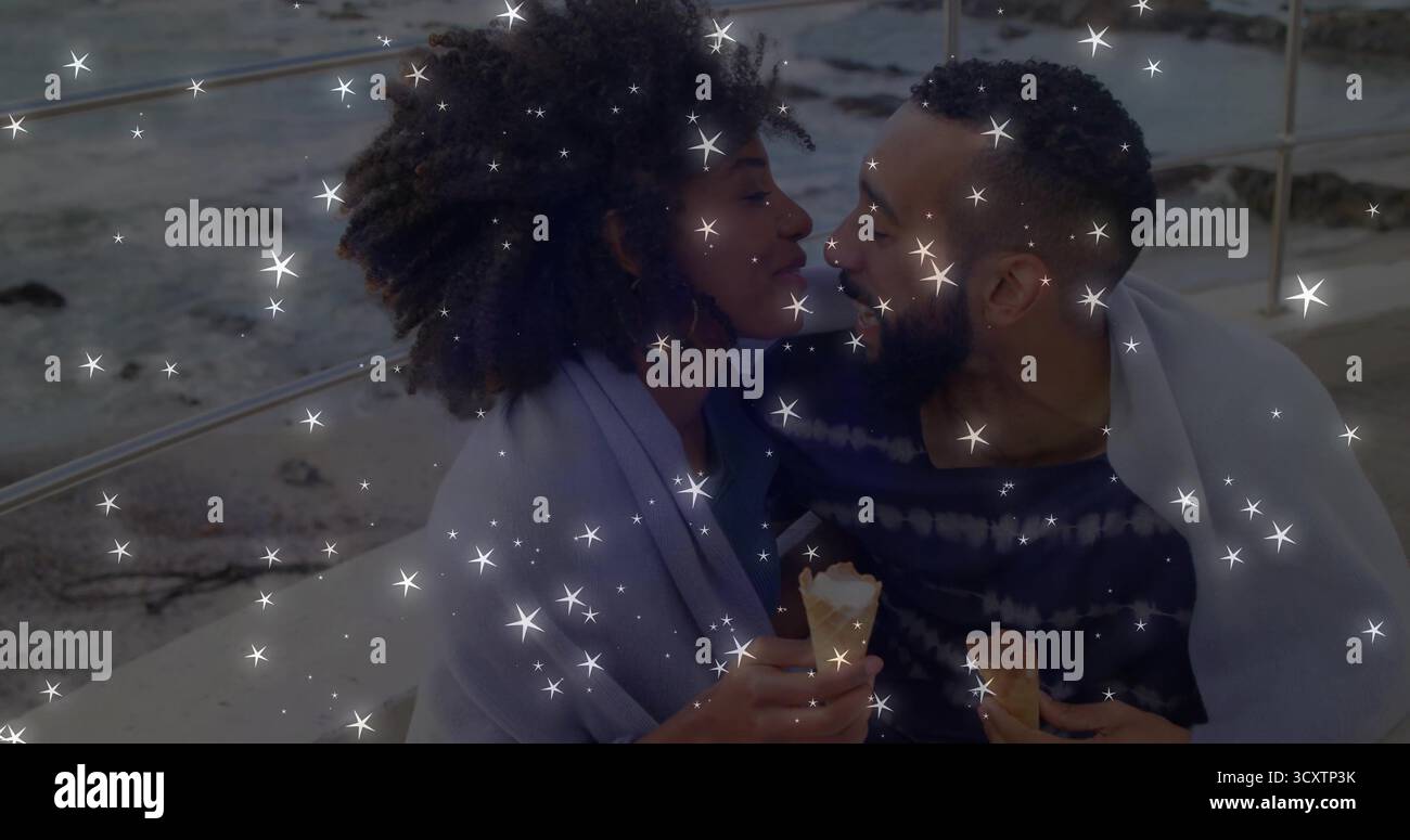 Coppia pendente che tiene in mano coni di vaniglia che si fondono sulla sporgenza dal mare, avvolta in coperta vicino a ringhiere metalliche Foto Stock