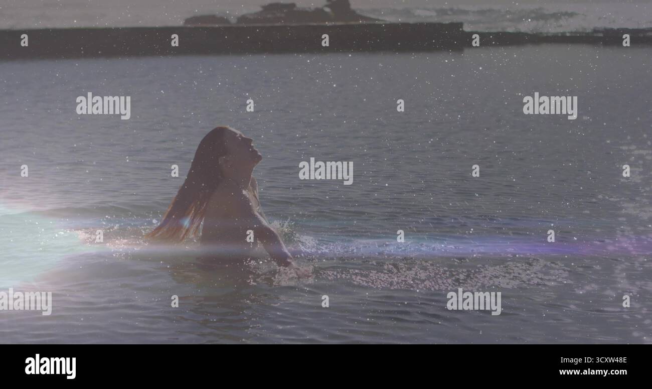 Nuotatore emergente che ribalta i capelli bagnati sul lago all'alba, con la parte superiore del costume da bagno, l'acqua aperta, il bagliore delle lenti Foto Stock