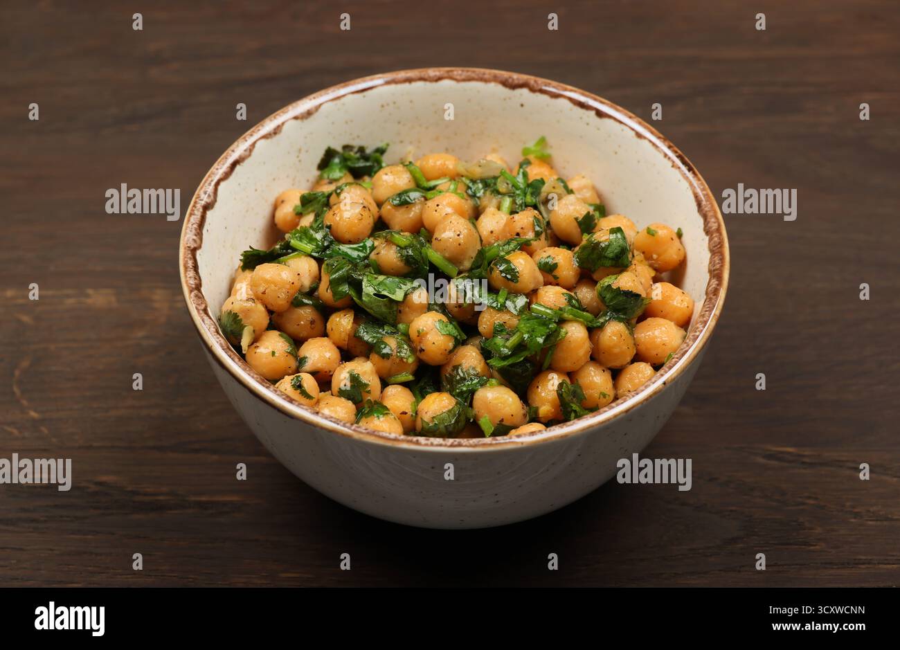 Ciotola in ceramica di ceci stagionati cotti con prezzemolo tritato e erbe verdi di cipolla si appoggia su un tavolo di legno marrone scuro, offrendo un sano pasto vegano Foto Stock