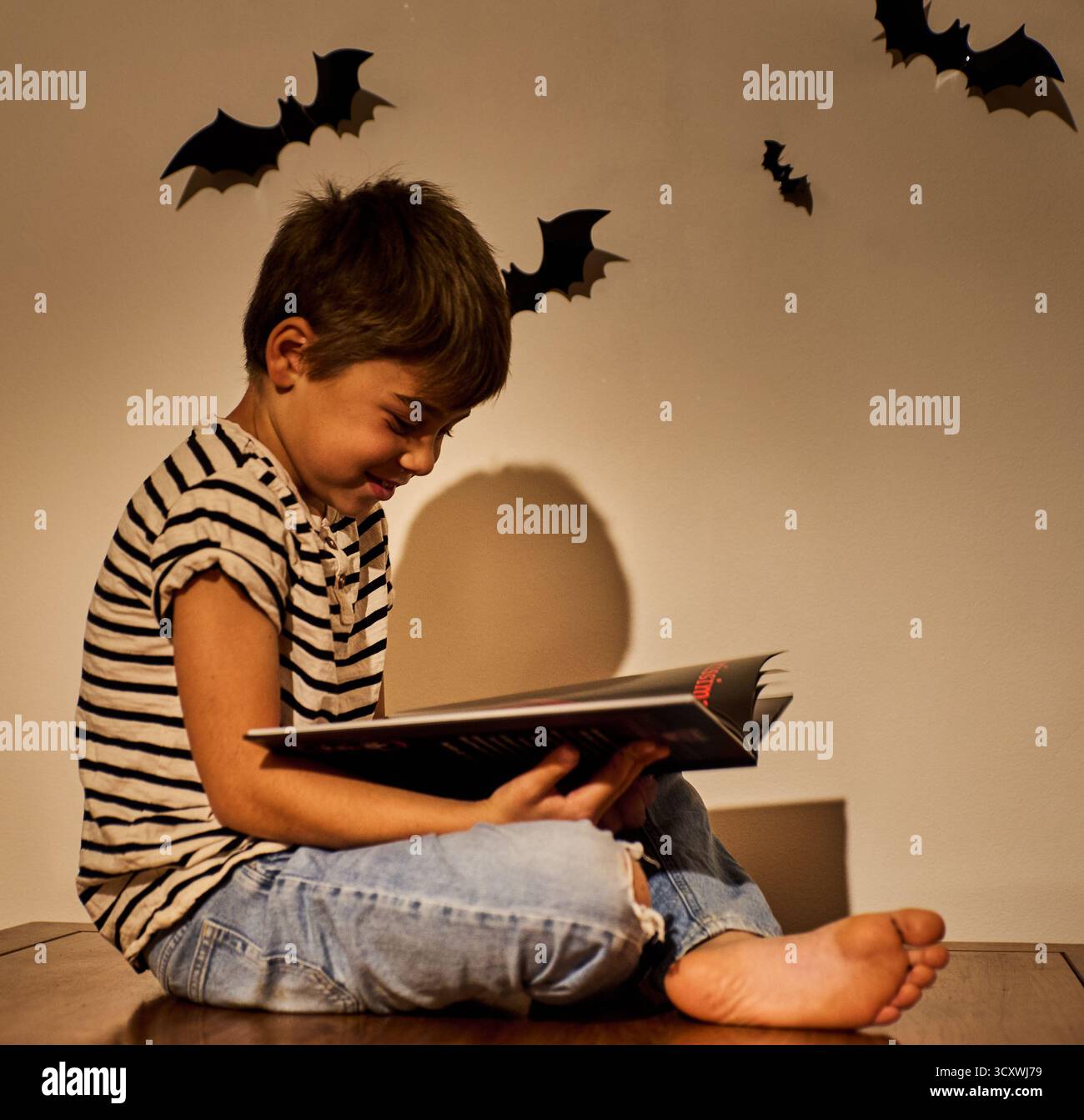 Bambino entusiasta o ragazzo sorridente che legge un libro nell'accogliente ambiente di Halloween con decorazioni di pipistrelli sulle pareti. L'illuminazione calda crea un autunno festoso e spettrale Foto Stock