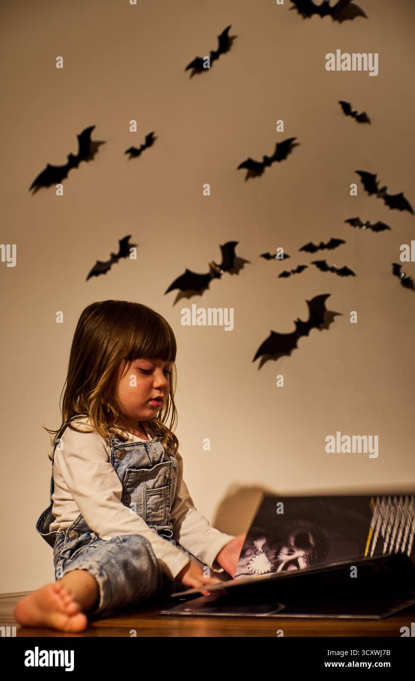 Un bambino affascinante o una ragazza seria in tuta legge un libro di Halloween sotto una luce calda, con decorazioni a parete per pipistrelli che creano un'atmosfera accogliente e spettrale Foto Stock