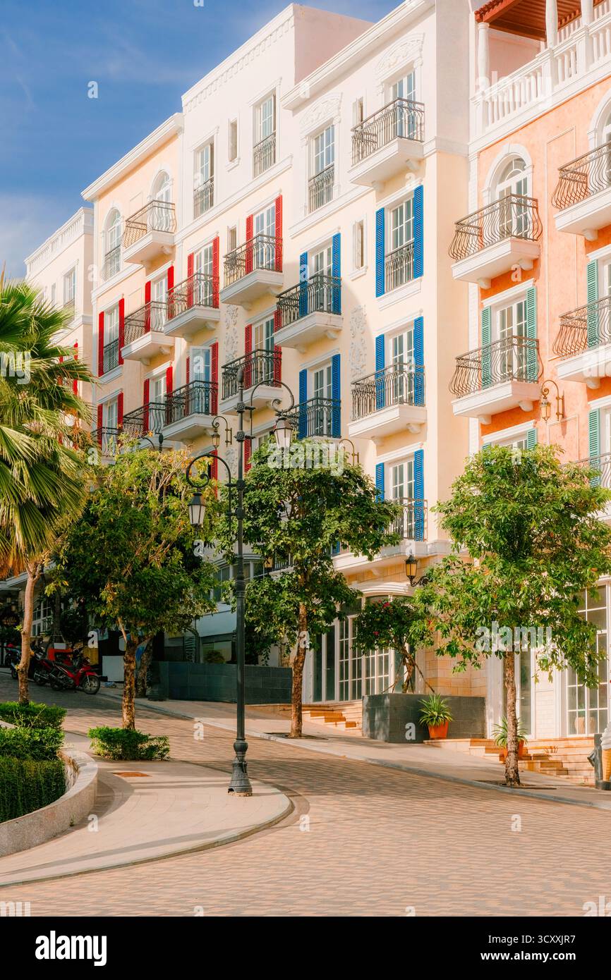 Colorati edifici in stile mediterraneo con balconi decorativi e luminose persiane fiancheggiano una strada tranquilla circondata da lussureggianti alberi verdi Foto Stock