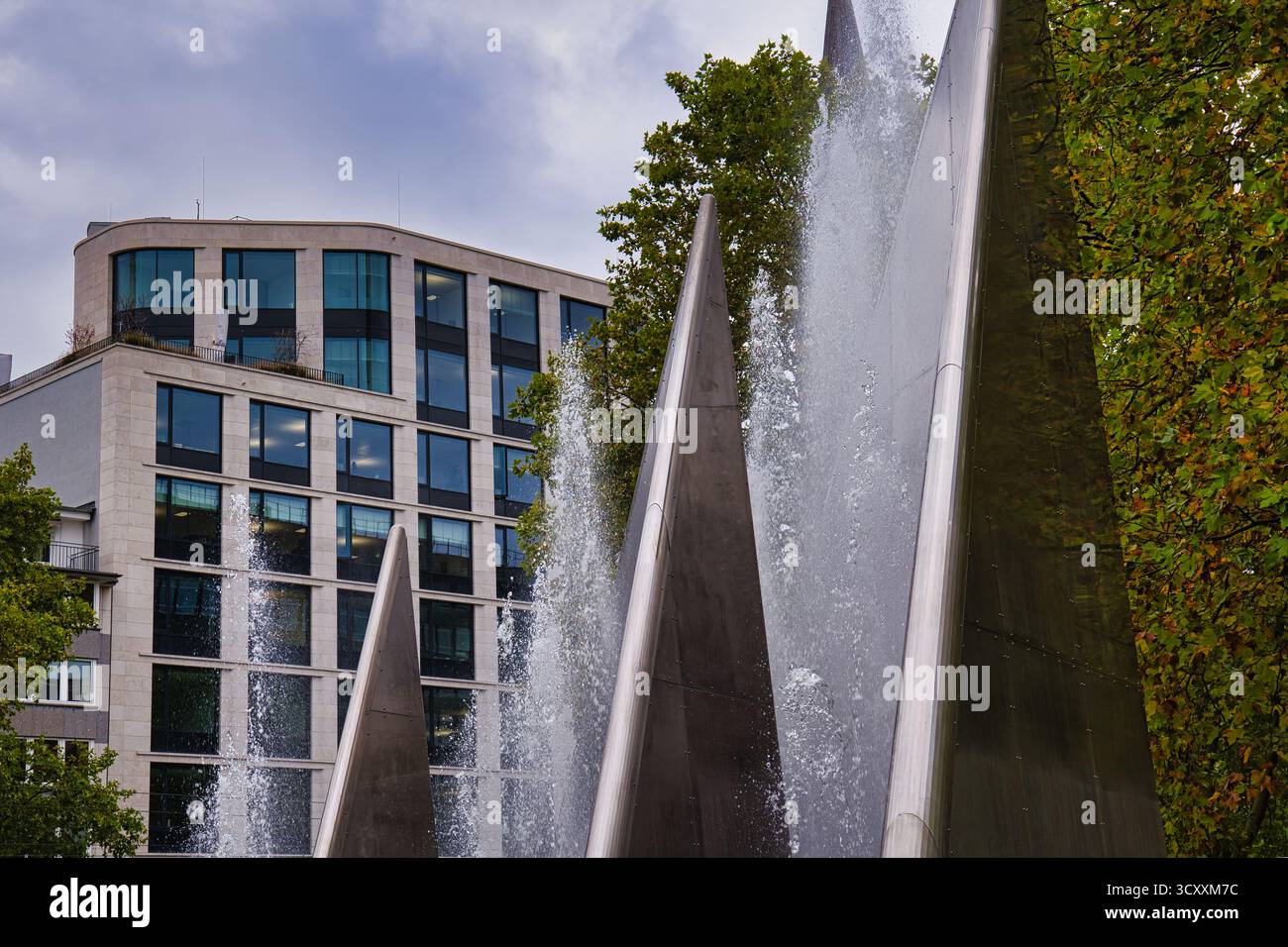 Fontana dinamica con sfondo architettonico moderno e vegetazione verde nel centro città Foto Stock