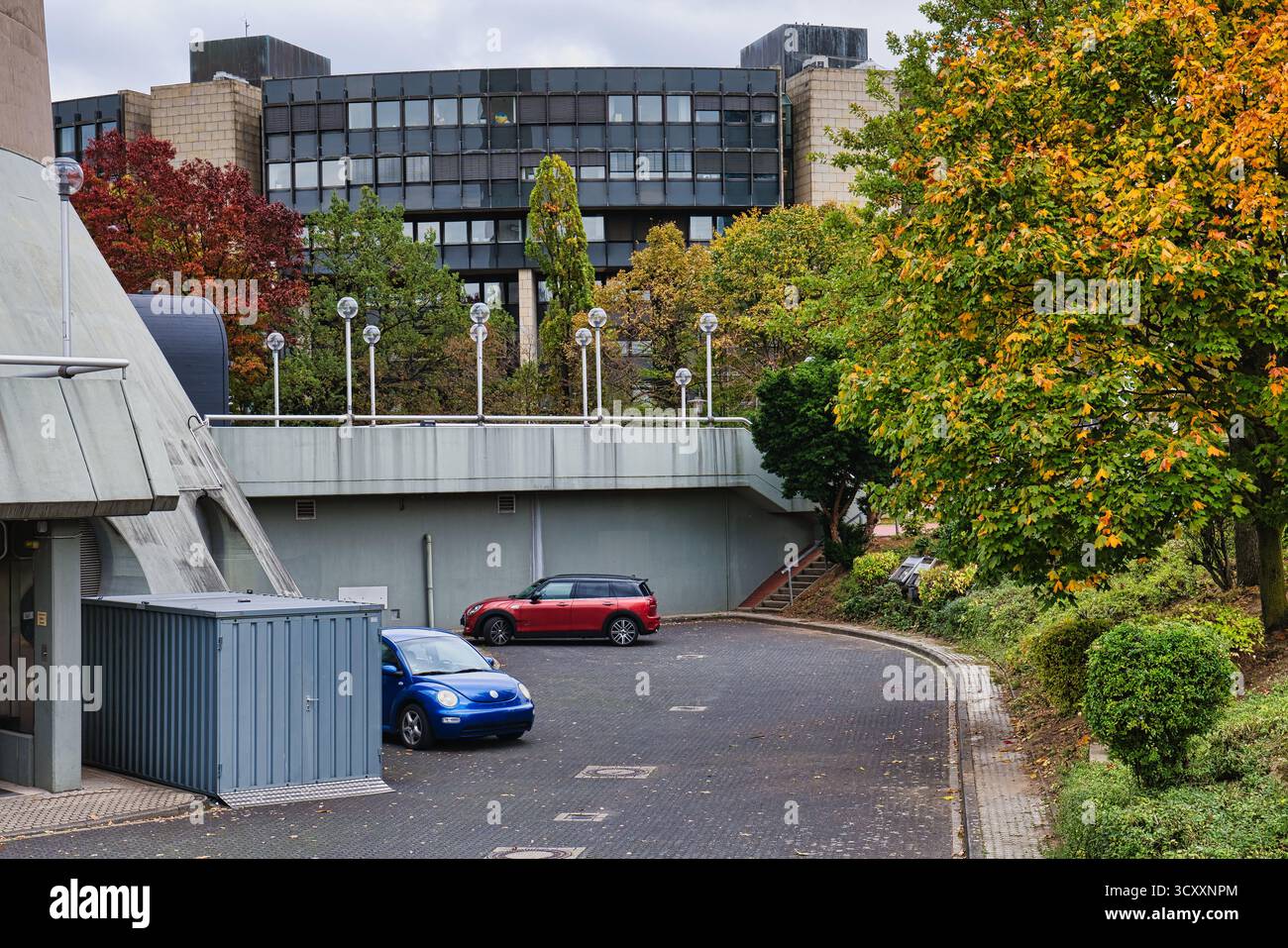 Rampa di parcheggio curva con scarabeo Volkswagen blu e SUV rosso parcheggiati sotto il ponte Gray Overpass, circondato da vibranti alberi autunnali e moderni edifici Foto Stock