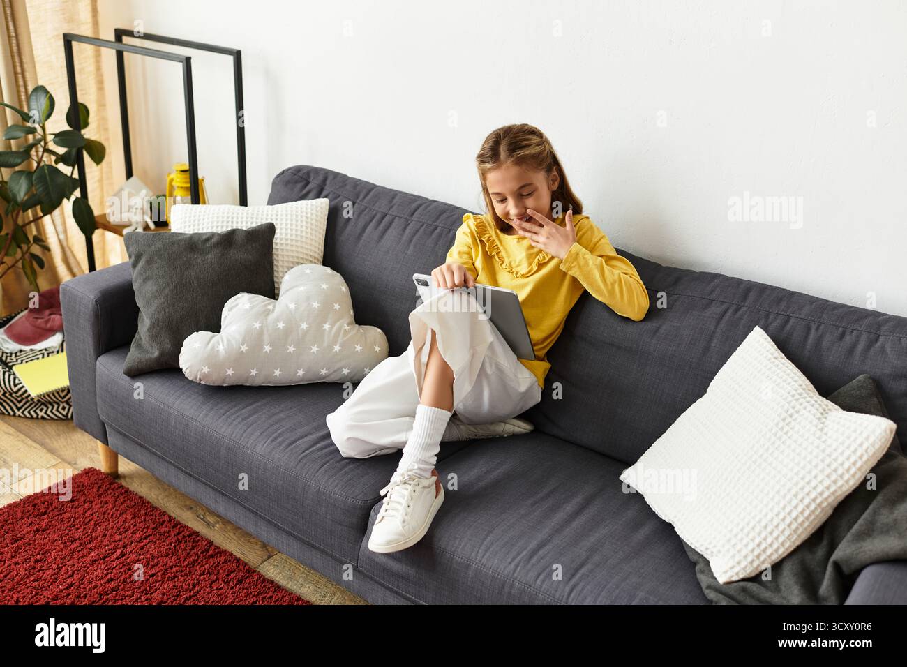 Una ragazza alfa di generazione in un outfit casual esplora felicemente il suo tablet mentre si rilassa a casa. Foto Stock