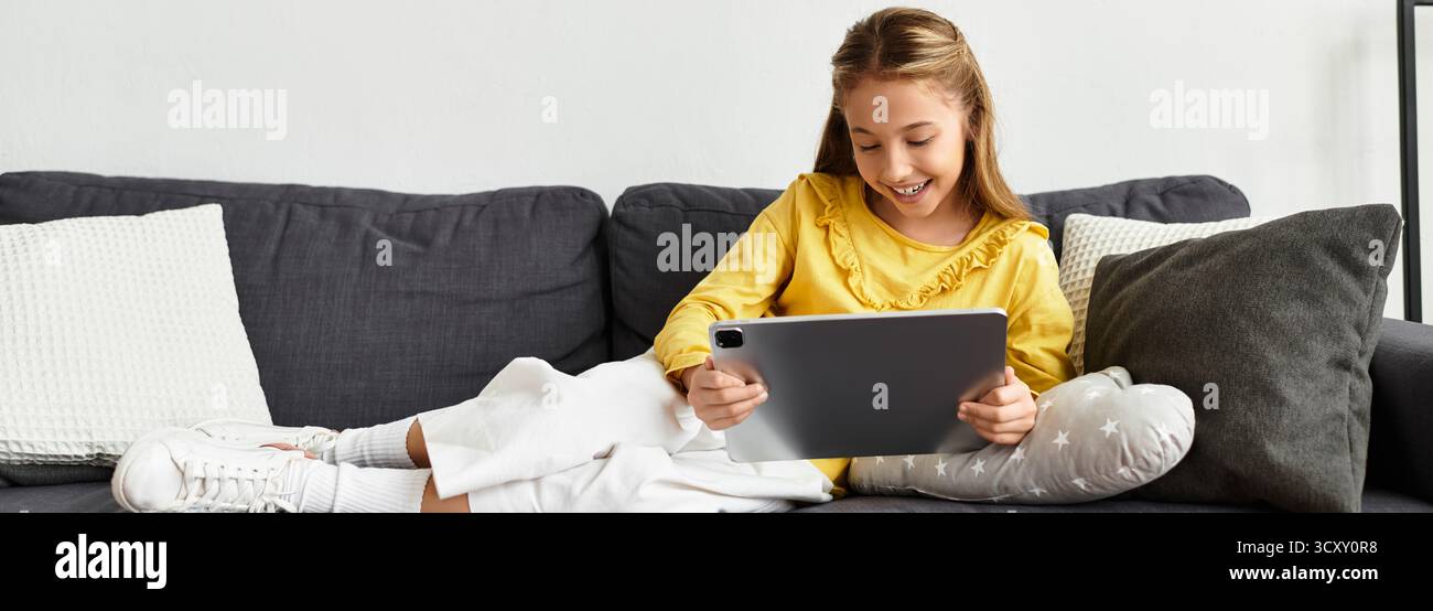 Una ragazza alfa di generazione in abiti casual sorride mentre si impegna con il suo tablet sul divano, sul banner Foto Stock