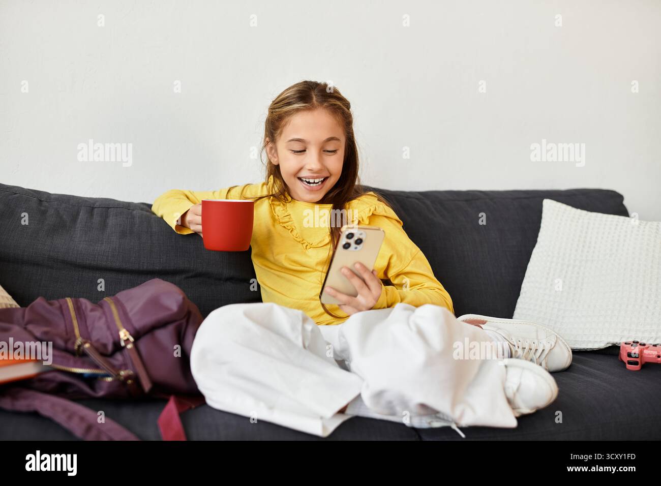 Un momento luminoso di una ragazza rilassata a casa con il suo smartphone e una bevanda calda in mano. Foto Stock
