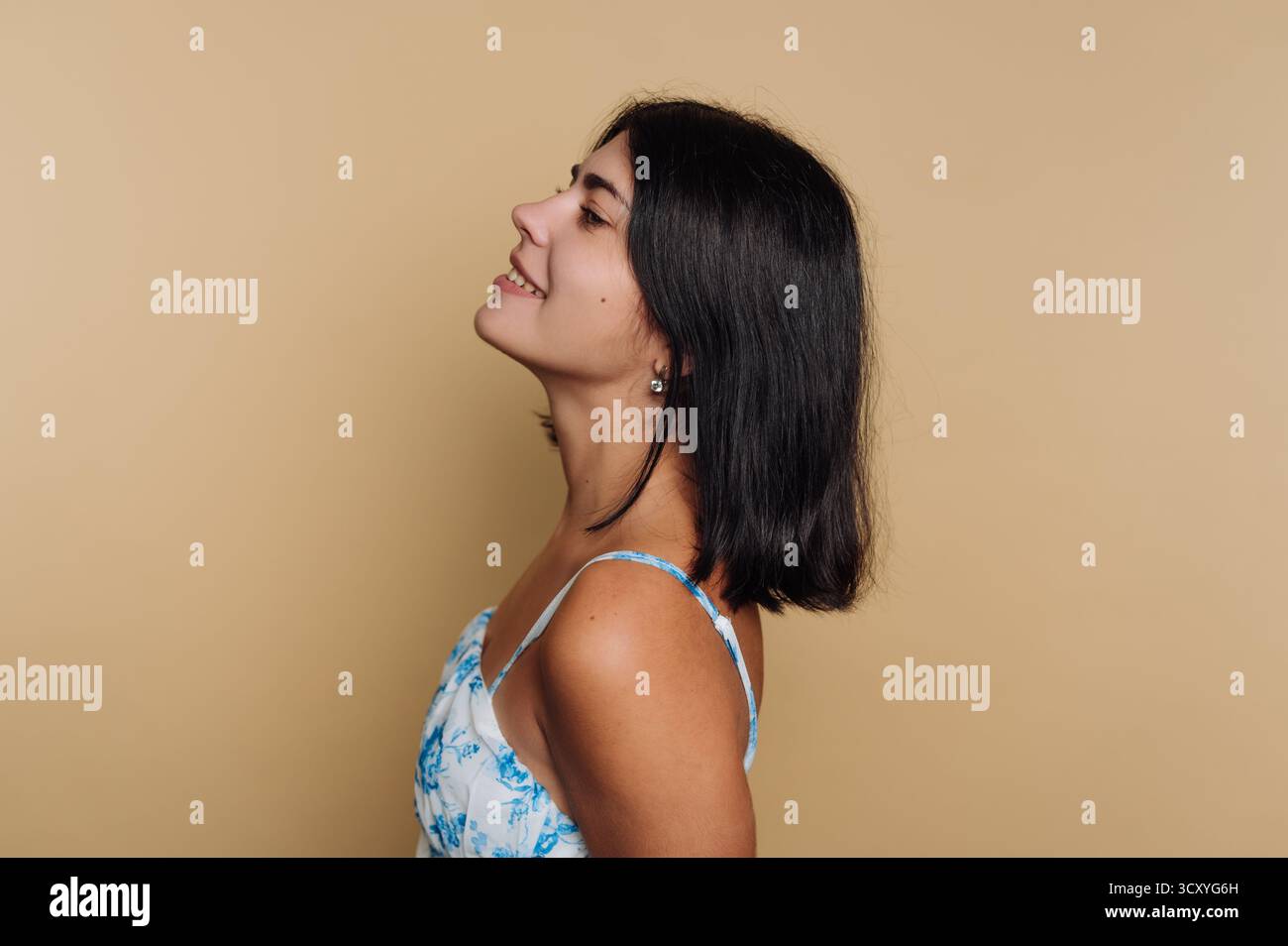 Una donna con i capelli scuri delle spalle sorride brillantemente mentre guarda di lato. Indossa un abito floreale azzurro e si erge di fronte a un essere morbido Foto Stock Una donna con i capelli scuri delle spalle sorride brillantemente mentre guarda di lato. Indossa un abito floreale azzurro e si erge di fronte a un essere morbido Foto Stock
