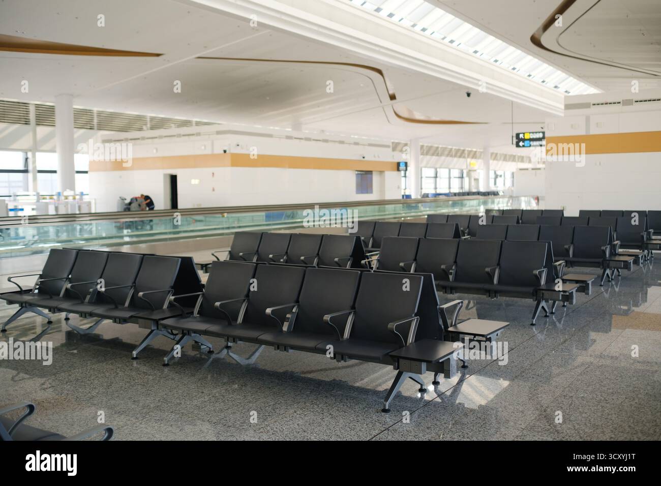 Una grande area salotto aperta in un moderno terminal aeroportuale presenta file di sedie nere disposte in modo ordinato. La luce del sole illumina lo spazio, evidenzia Foto Stock