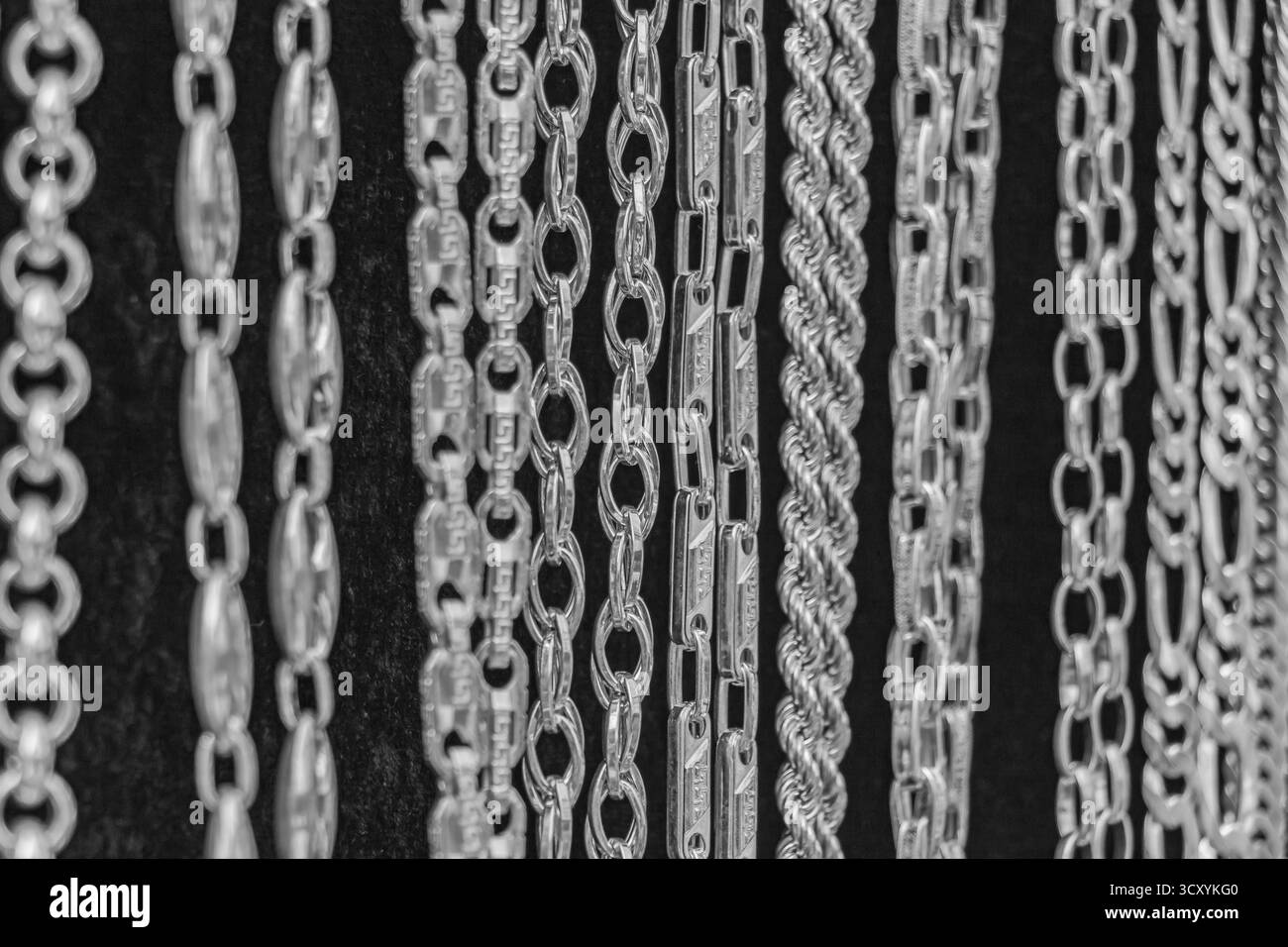 Primo piano monocromatico di varie catene d'argento appese in file su sfondo scuro. Ogni pezzo mostra una raffinata maestria artigianale e un motivo a maglie intricate Foto Stock