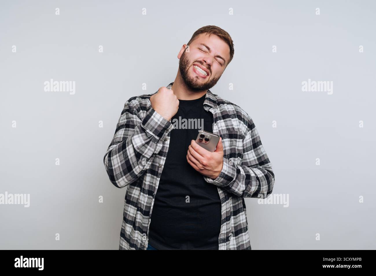 Un uomo sta in casa, sorridendo e stringendo il pugno con entusiasmo mentre tiene in mano uno smartphone. Indossa una camicia a quadri e sembra che si stia godendo un deposito Foto Stock