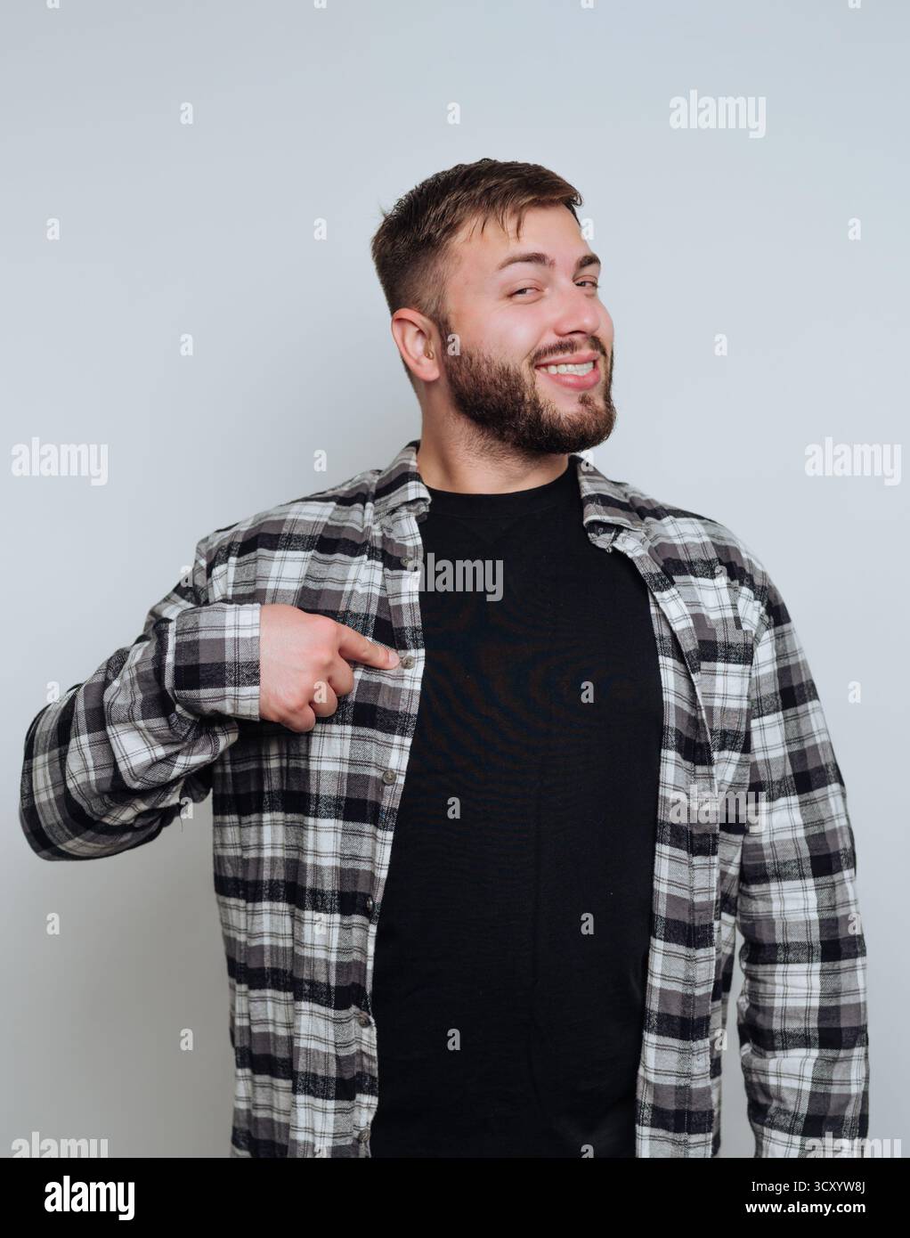 Un giovane uomo si trova in una stanza ben illuminata con un sorriso allegro e indossa una camicia nera sotto una camicia aperta a quadri. Indica con sicurezza il suo petto, espr Foto Stock