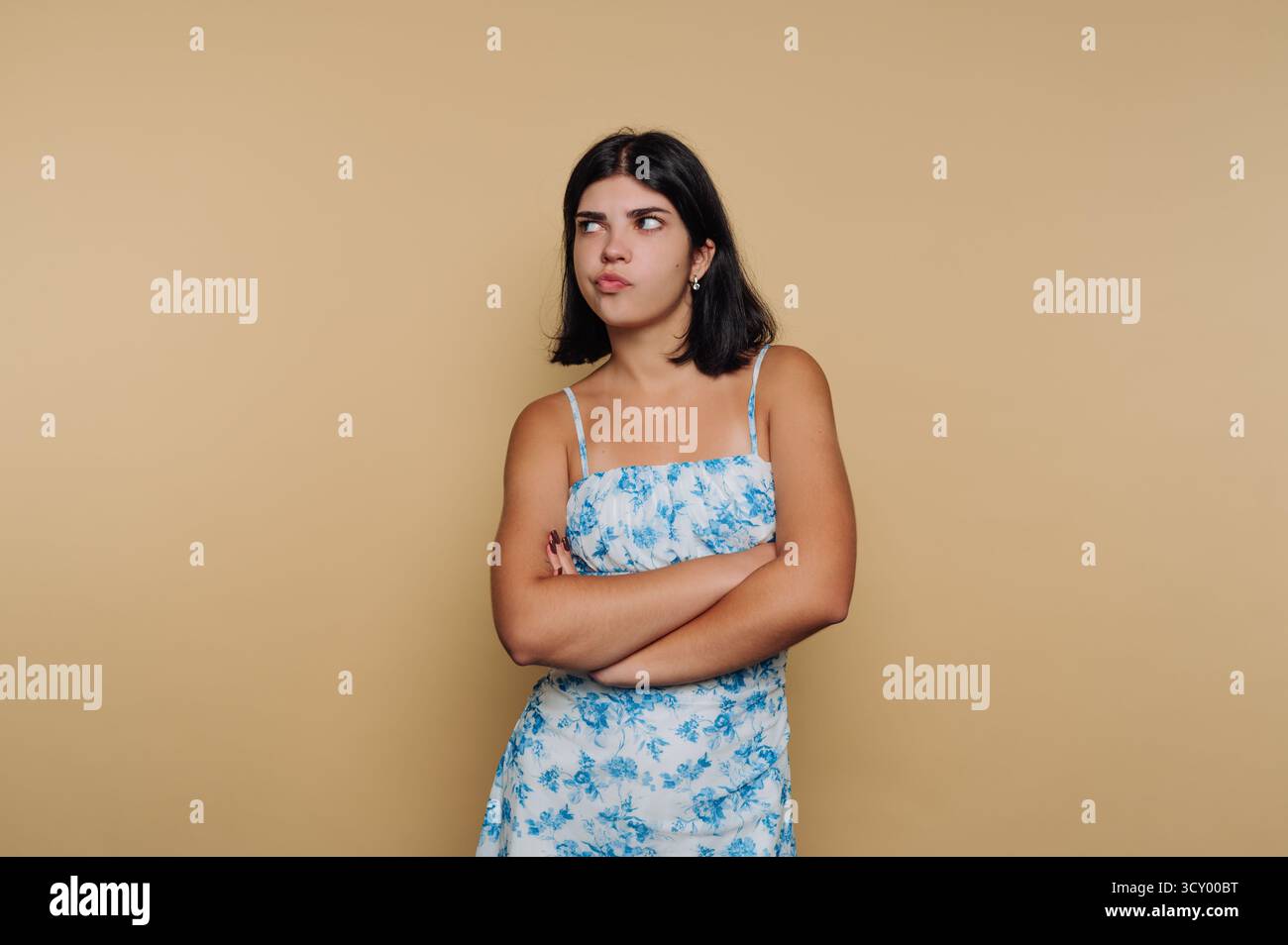 Una giovane donna sta in piedi con le braccia incrociate, mostrando un aspetto scettico. Indossa un abito floreale ed è in una stanza semplice con uno sfondo di colore neutro Foto Stock