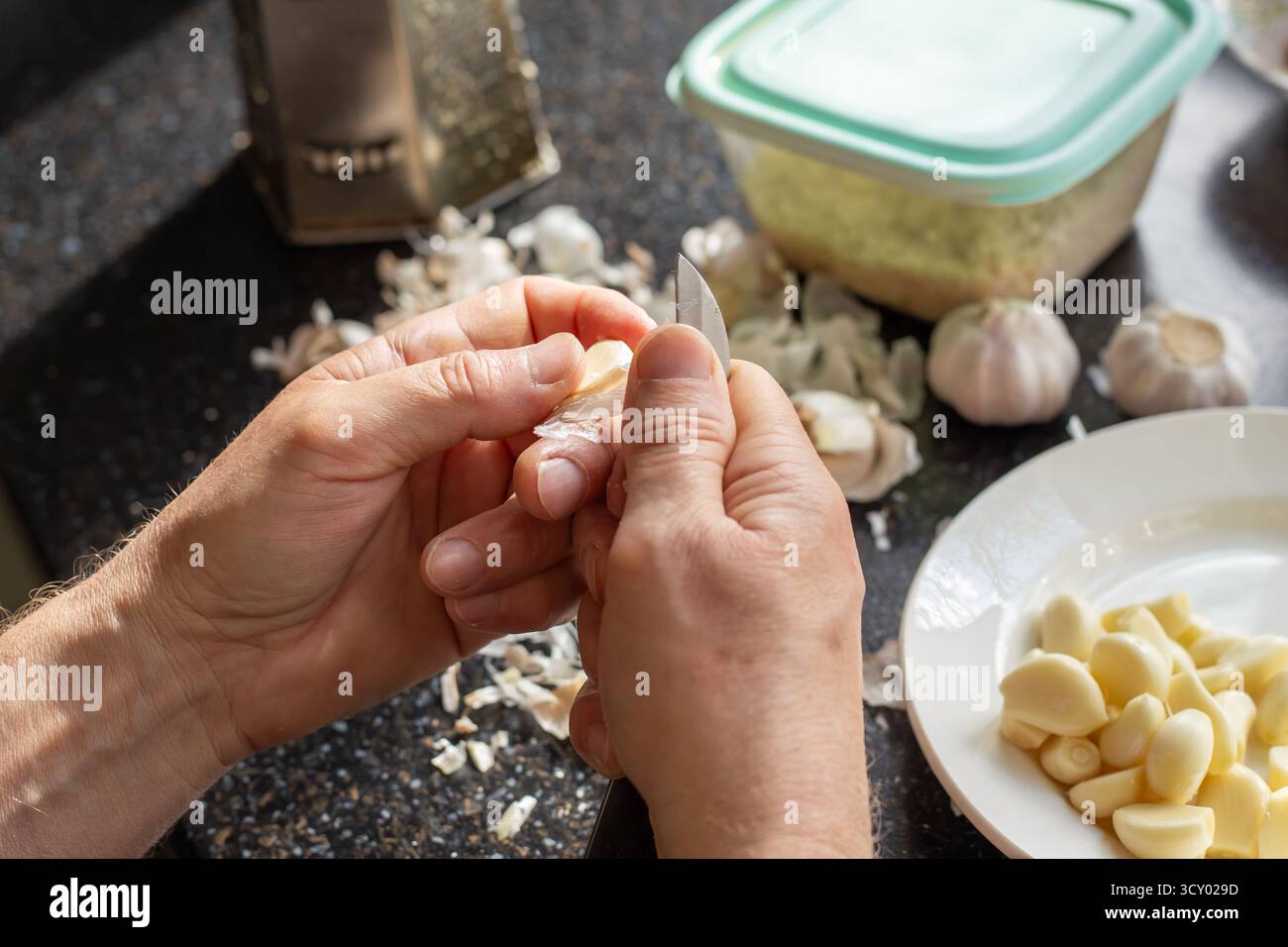 Un uomo sbuccia gli spicchi d'aglio con un coltello da cucina, preparando l'ingrediente per la cottura su un piano di lavoro. Foto Stock