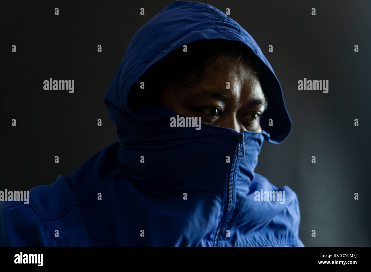 Uomo indonesiano anni '30 con barba e giacca in nylon blu, testa e parte del viso oscurati dal cappuccio, suggestivo ritratto urbano con sfondo nero Foto Stock