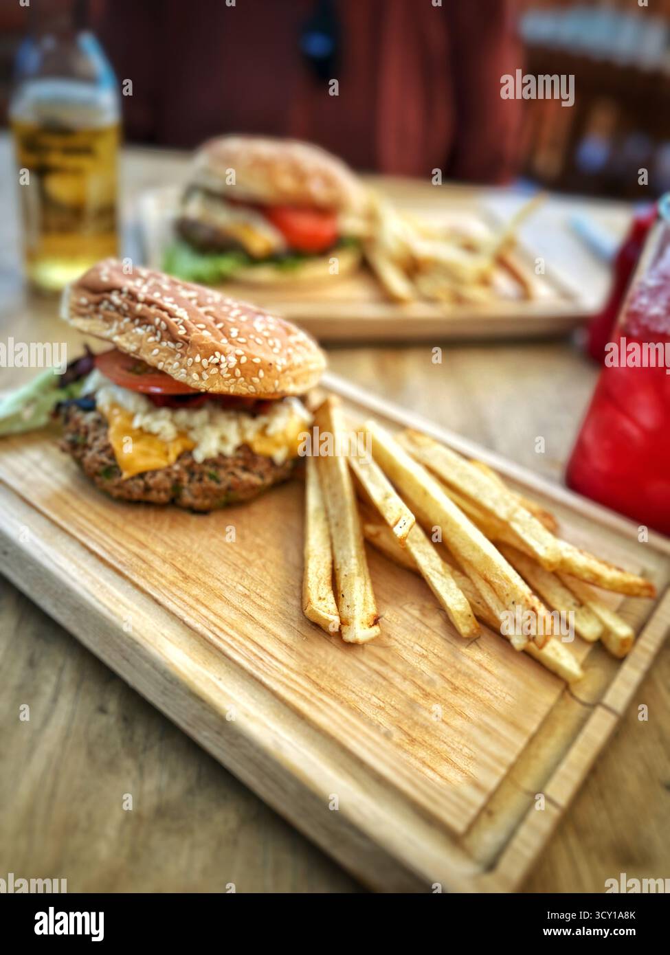 Due hamburger con patatine fritte serviti su tavole di legno su un tavolo insieme a bevande rinfrescanti in un ambiente informale Foto Stock