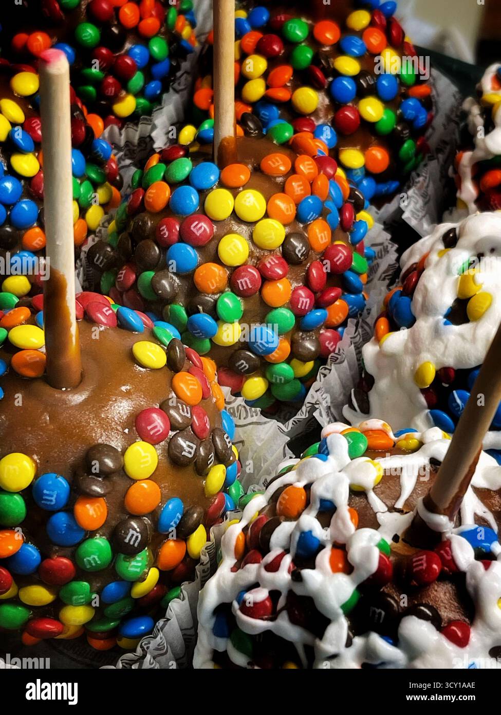 Primo piano di mele al caramello ricoperte di cioccolatini colorati ricoperti di caramelle e glassa bianca, esposte in bicchieri di carta decorativi - Immagine stock catturata con smartphone