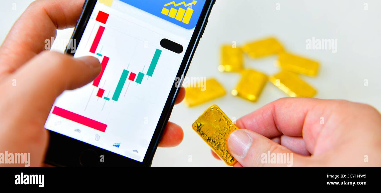 Tenere a mano uno smartphone e una barra d'oro per controllare il grafico del prezzo dell'oro per l'investimento Foto Stock