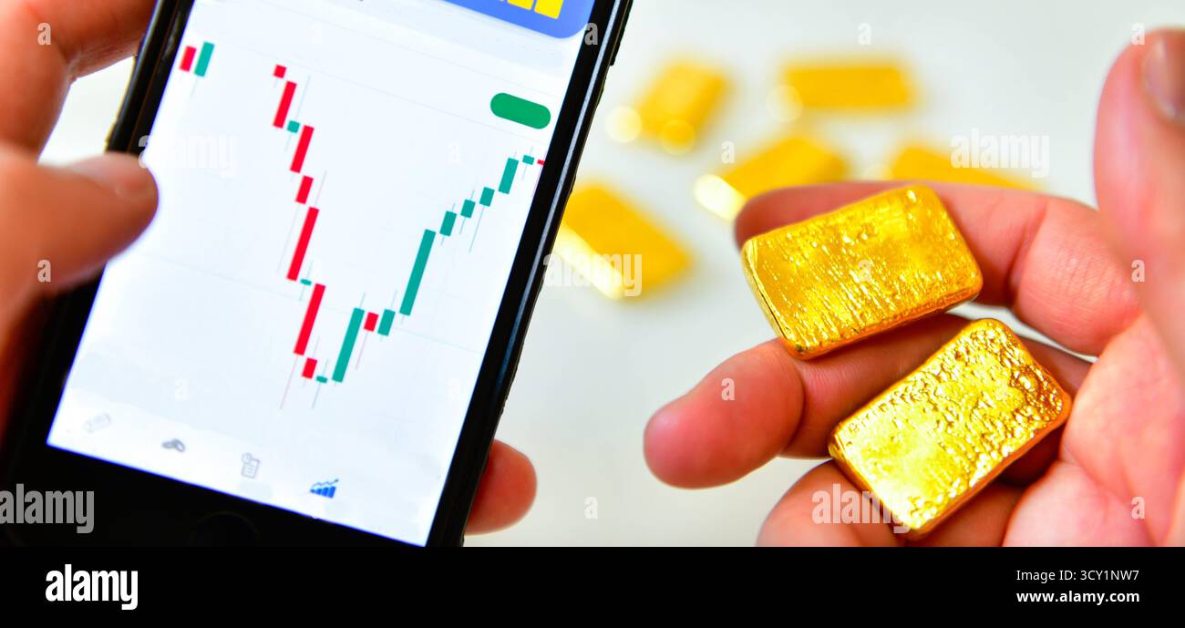 Tenere a mano uno smartphone e una barra d'oro per controllare il grafico del prezzo dell'oro per l'investimento Foto Stock