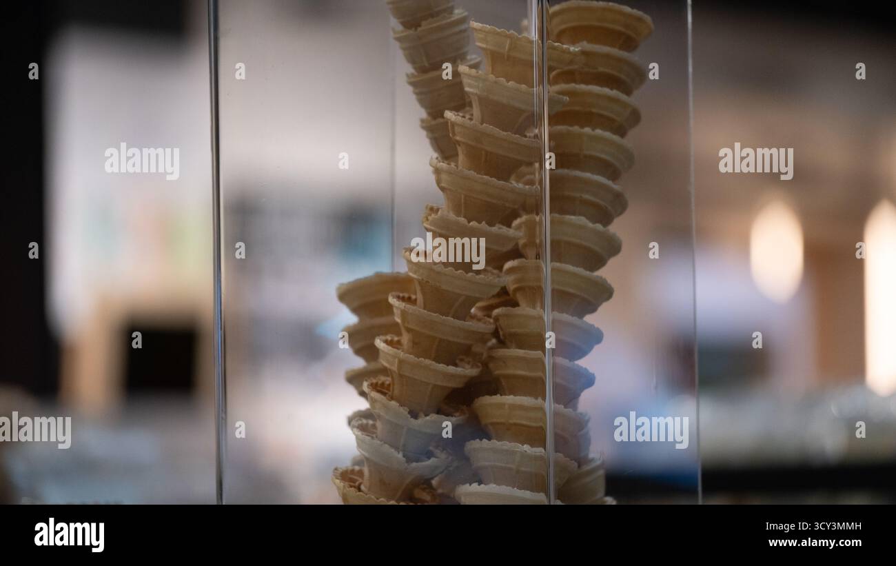 Tazze di gelato con cialde impilate esposte in un contenitore di vetro trasparente all'interno di un moderno caffè. Il caldo bokeh e l'illuminazione soffusa creano un ambiente intimo e minimalista Foto Stock