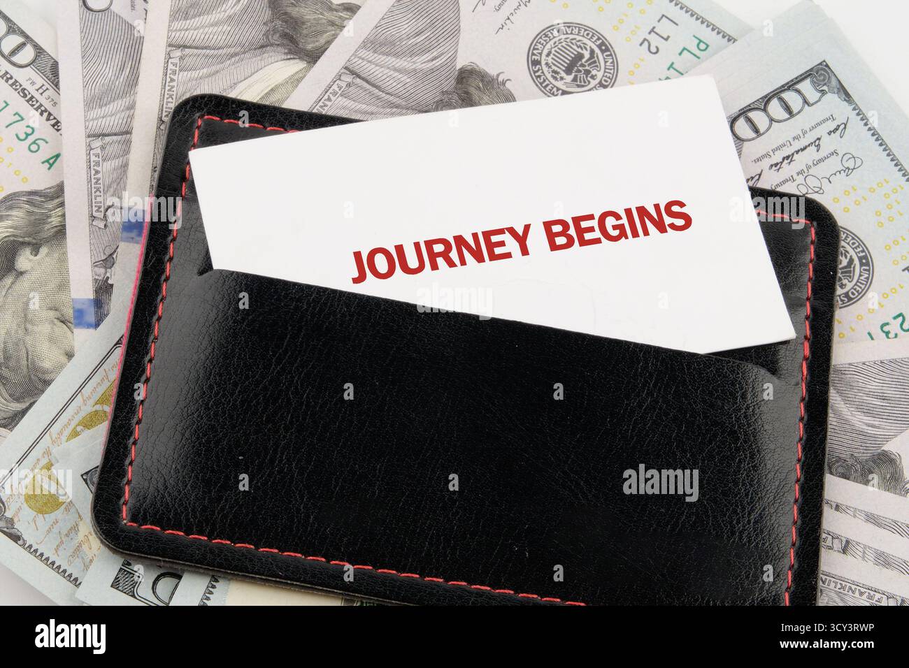Un elegante portafoglio nero contiene una carta che dice che il viaggio inizia, circondata da banconote da cento dollari, a simboleggiare l'inizio di un'entusiasmante attività finanziaria Foto Stock