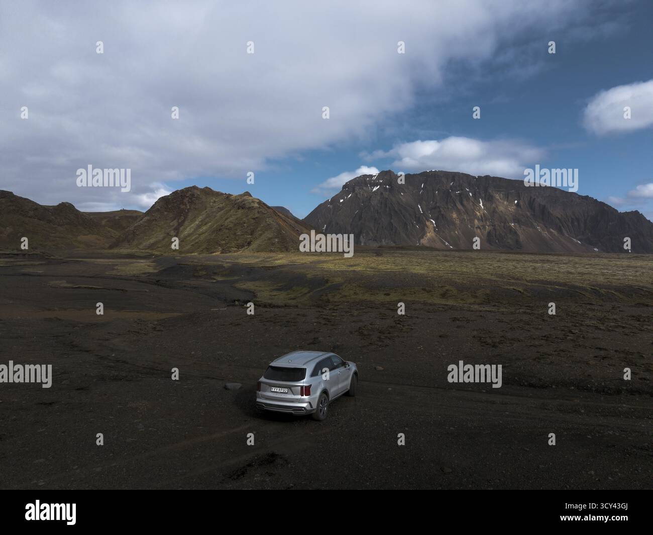 Veduta aerea di un SUV solitario parcheggiato sulla terra vulcanica nera, sullo sfondo di aspre montagne sotto un vasto cielo, Reykjavík, Islanda. Foto Stock