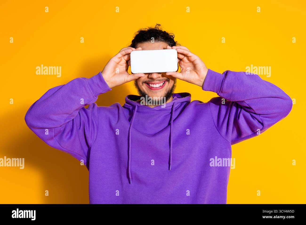 Uomo sorridente che posa su sfondo giallo con il telefono in mano con cappuccio viola per promuovere lo stile di vita moderno e il divertimento Foto Stock