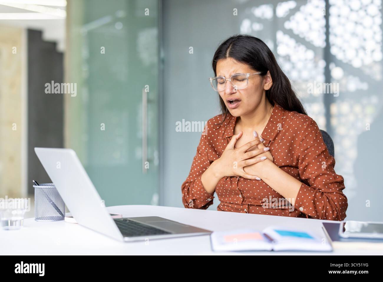 Giovane donna indiana che soffre improvvisamente di gravi dolori al petto o problemi cardiaci mentre è seduta alla sua scrivania, tiene il petto con entrambe le mani ed esprime disagio e sofferenza Foto Stock
