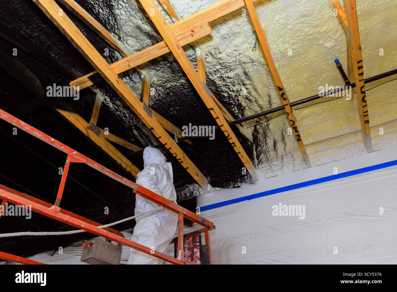 L'appaltatore di dispositivi di protezione applica l'isolamento della vernice refrattaria utilizzando una pistola a spruzzo alle travi in legno del soffitto nel cantiere. Foto Stock