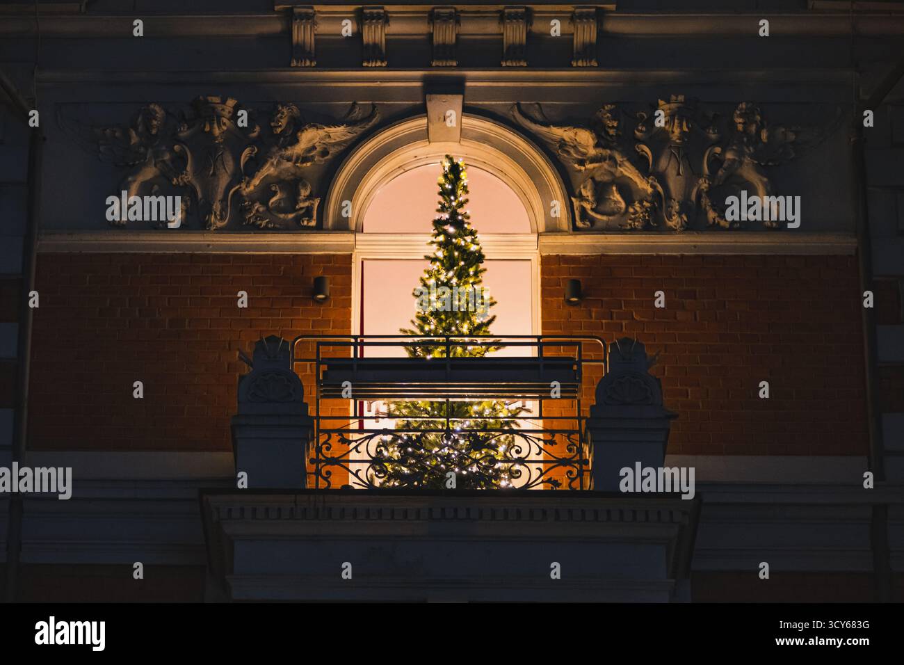 Un albero di Natale illuminato si illumina calorosamente dietro un balcone ornato, incorniciato da intricati dettagli architettonici e muratura nel quartiere della città Vecchia Foto Stock