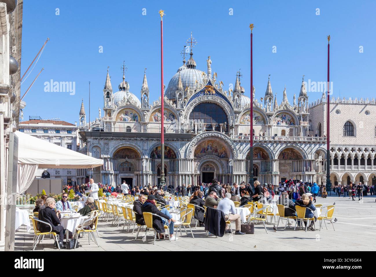 Turisti che si rilassano in primavera al sole nel ristorante all'aperto di fronte a Basilica San Marco, Piazza San Marco, San Marco, Venezia, Veneto, Italia Foto Stock