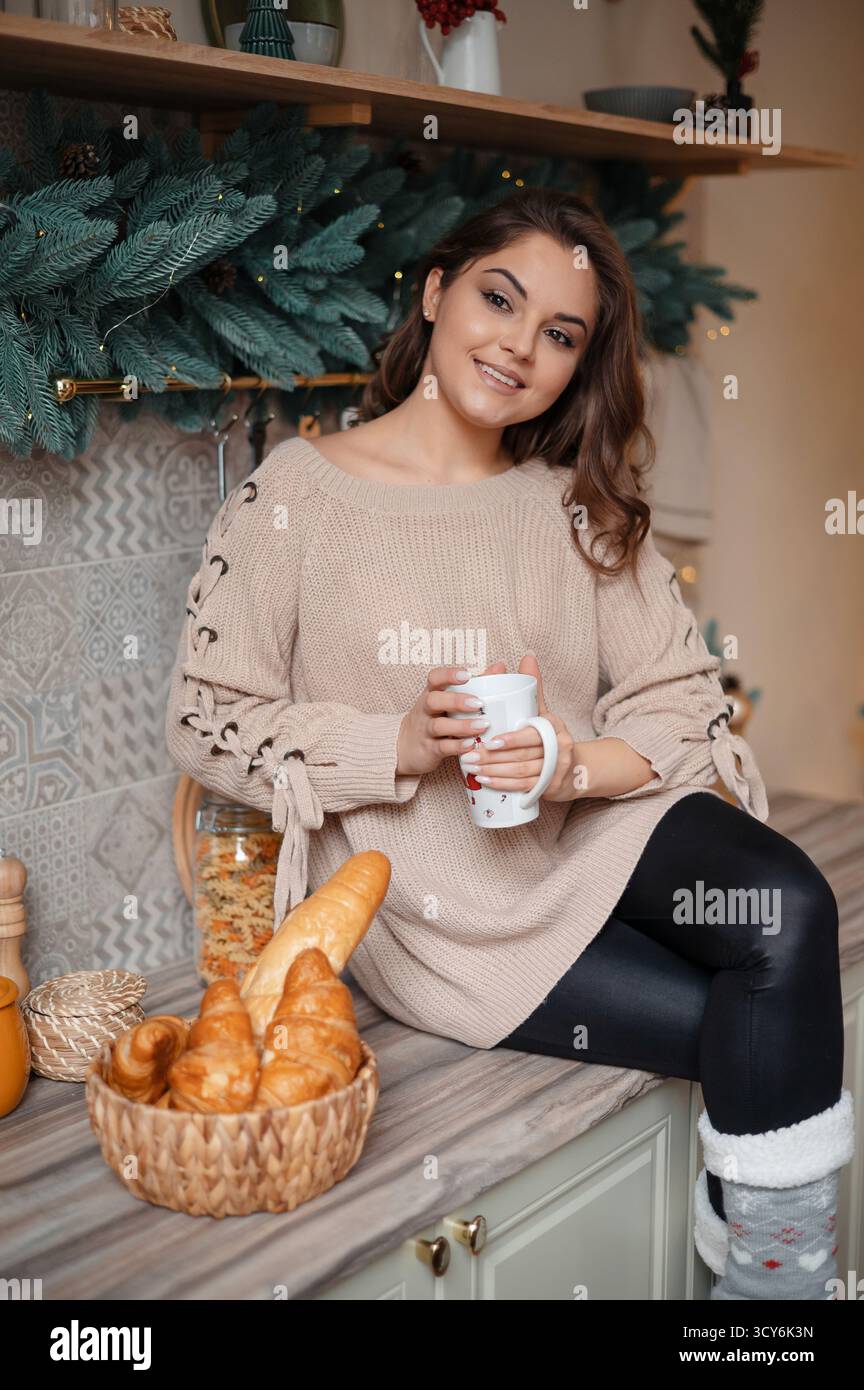 Donna in un comodo maglione che tiene in mano una tazza, seduta accanto a un cestino di dolci. Foto Stock