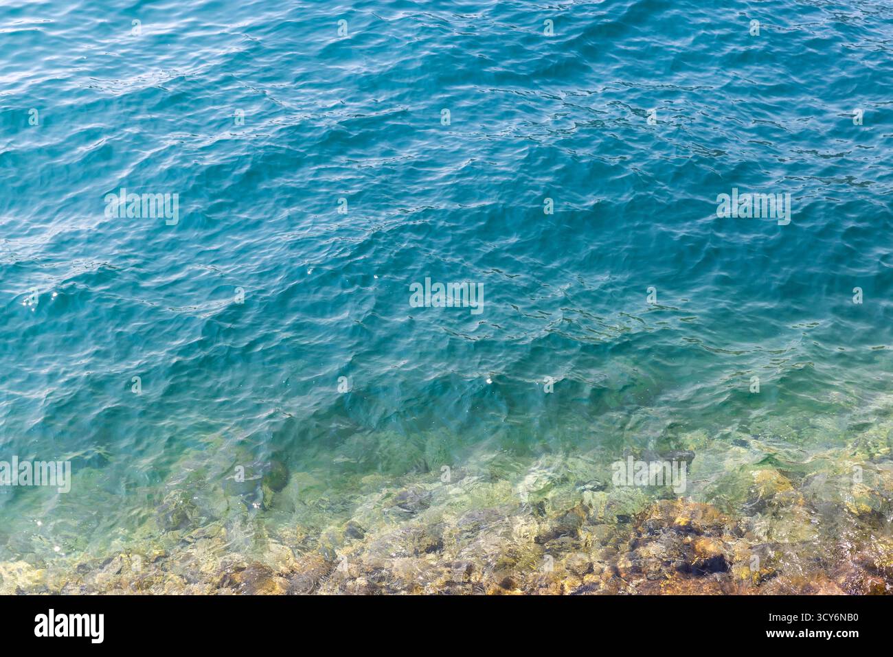 Il mare cristallino e turchese incontra una costa rocciosa, con onde dolci e ondulazioni illuminate dal sole. Una tranquilla scena costiera perfetta per viaggi, natura o relax Foto Stock