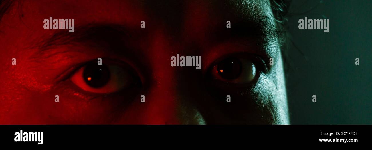 Primo piano dell'occhio dell'uomo indonesiano con luce ambientale rossa e verde, misteriosa espressione evidenziata su sfondo scuro nel drammatico studio portra Foto Stock