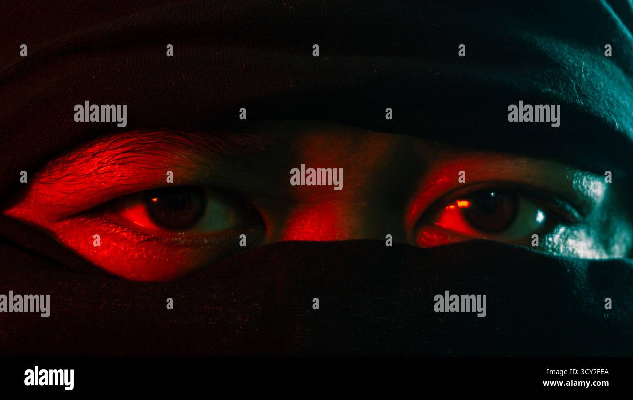 Primo piano di un occhio maschile che indossa una maschera sotto la luce ambientale rossa e verde, misterioso sguardo in stile ninja catturato su sfondo nero in un ritratto spettacolare Foto Stock
