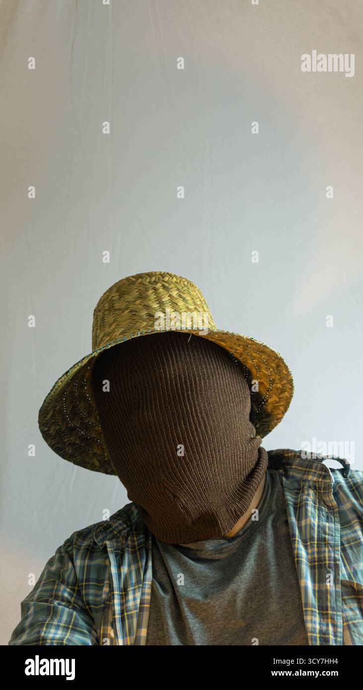 Primo piano di un agricoltore mascherato che indossa un cappello di paglia, viso completamente coperto di tessuto marrone, sfondo bianco che enfatizza il mistero e l'identità agricola Foto Stock