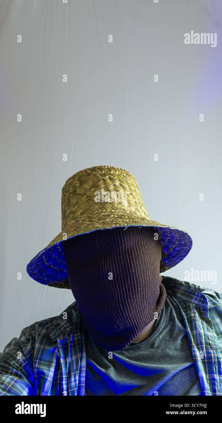 Primo piano di un agricoltore mascherato con cappello di paglia sotto la luce blu dell'ambiente, volto invisibile, carattere rurale con illuminazione stilizzata su sfondo bianco semplice Foto Stock