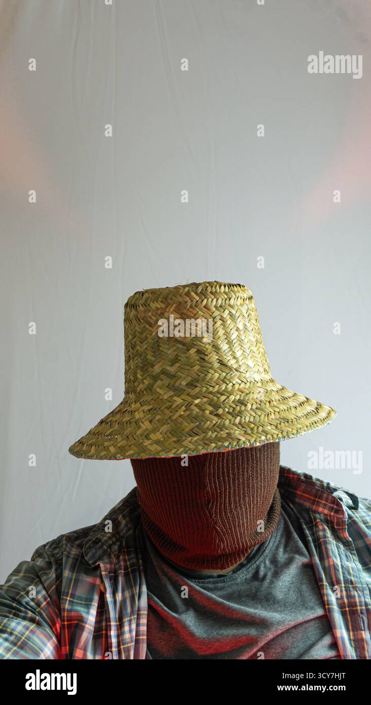 Contadino mascherato in cappello di paglia con volto nascosto, luce rossa aggiunge un tono drammatico al ritratto rurale, sfondo bianco minimalista con un'atmosfera intensa Foto Stock