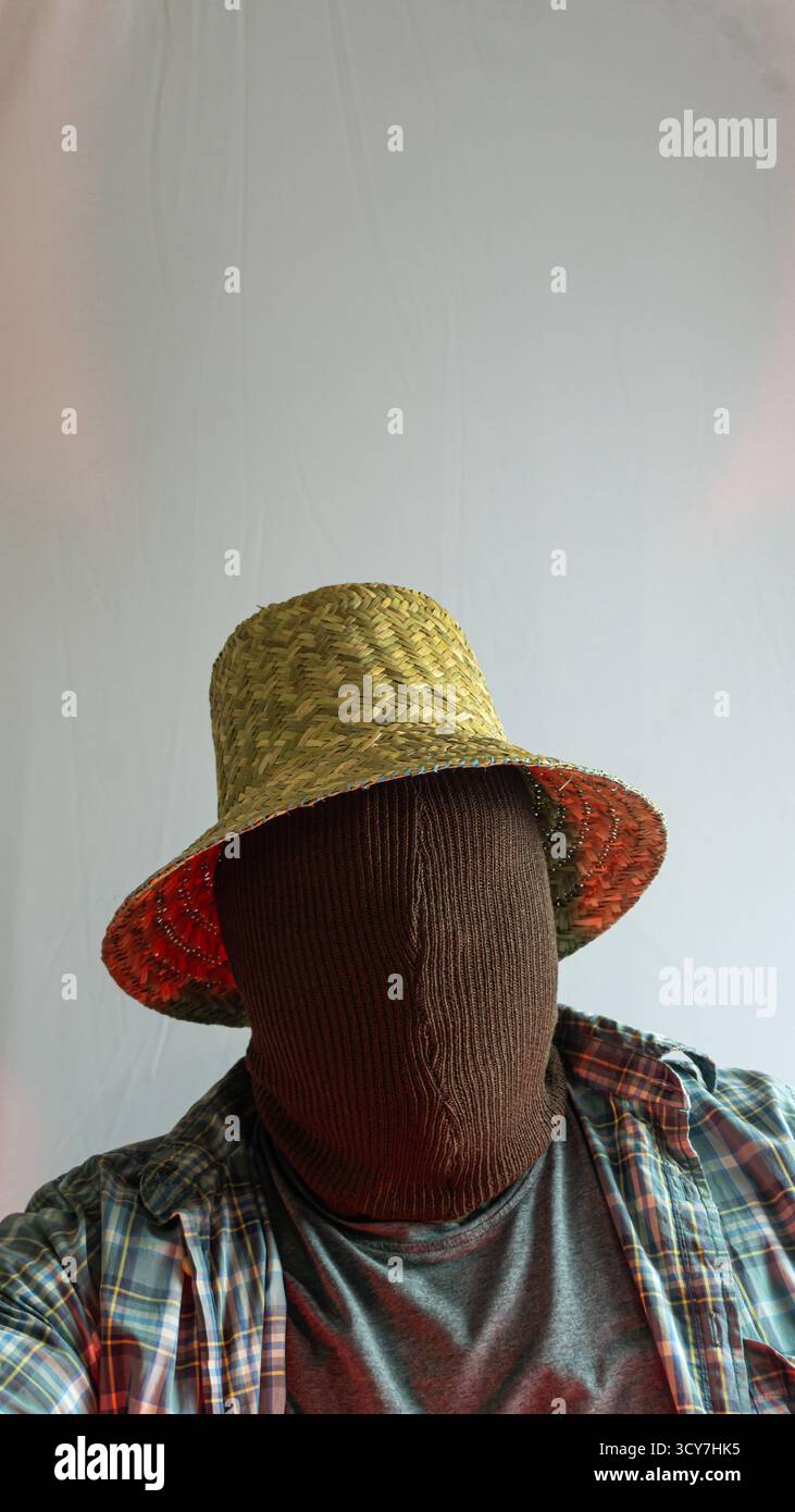 Contadino mascherato in cappello di paglia con volto nascosto, luce rossa aggiunge un tono drammatico al ritratto rurale, sfondo bianco minimalista con un'atmosfera intensa Foto Stock
