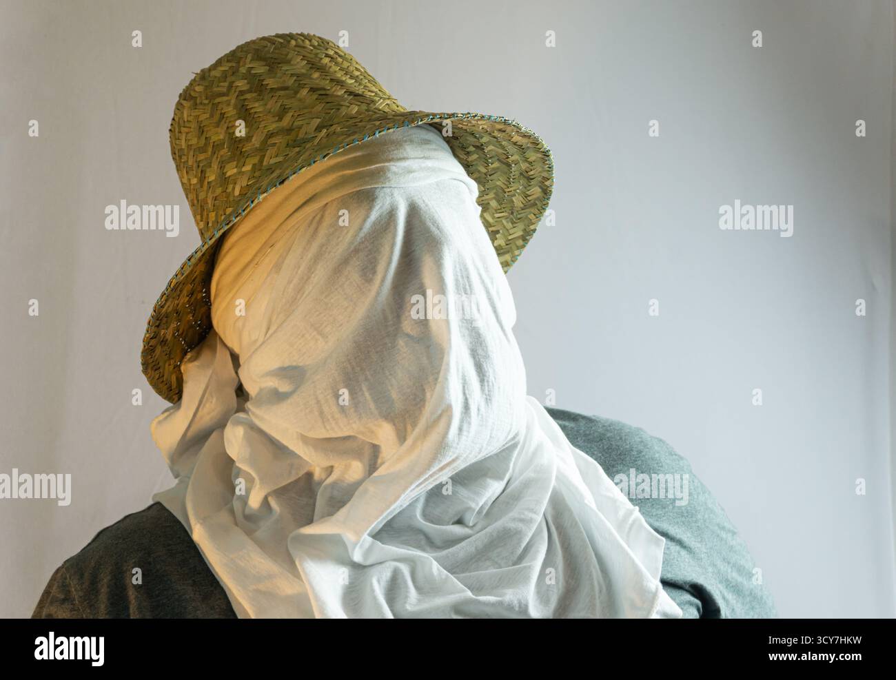 Primo piano di un agricoltore che indossa un cappello di paglia e una copertura facciale in tessuto bianco, anonima identità rurale mostrata su sfondo bianco pulito in un portrai minimalista Foto Stock